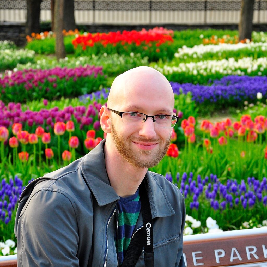 Photo of a man with light skin, a bald head, glasses, and a beard. He is sitting on a bench in a park, with pink and purple tulips in the background.